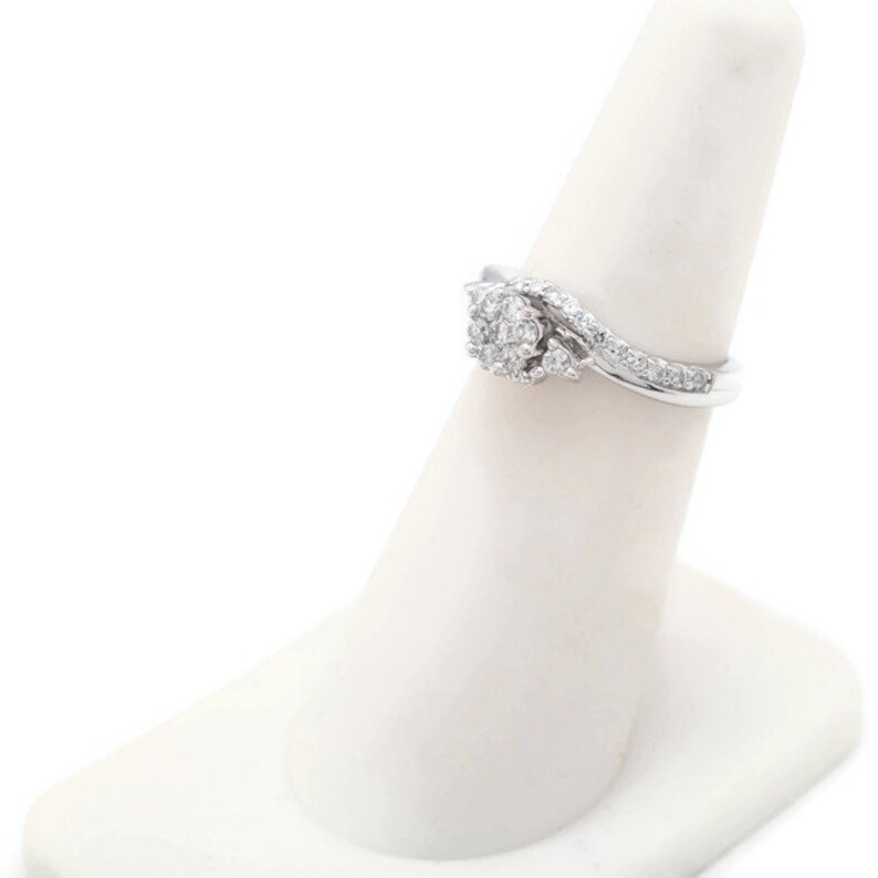 Op de afbeelding: Een zilveren ring met een cluster van kleine diamanten gezet in een swirl-ontwerp. De ring is op een witte ringstandaard.