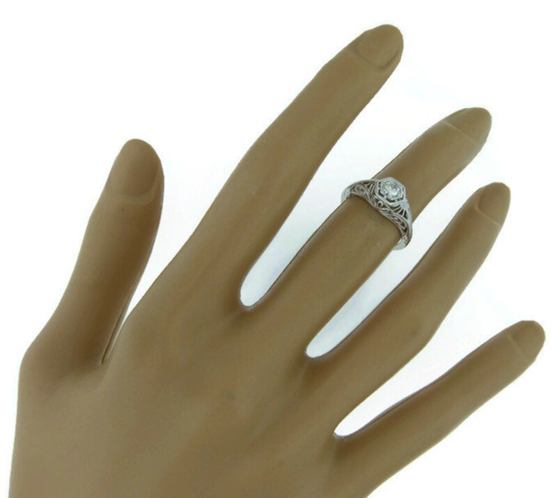 May include: A white gold engagement ring with a filigree design and a single round diamond in the center.