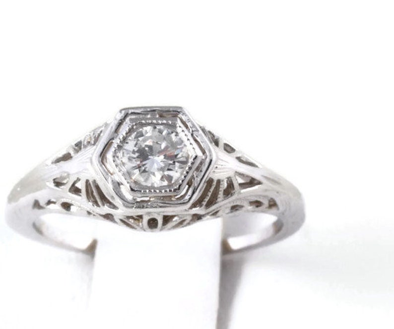 May include: A silver filigree ring with a single round diamond set in a hexagonal bezel.
