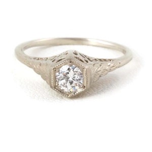May include: A silver engagement ring with a round, clear diamond set in a hexagonal bezel. The ring band is decorated with intricate filigree details.