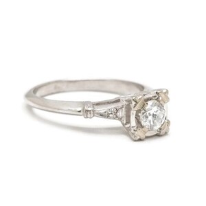 May include: A silver engagement ring with a single round diamond set in a square-shaped prong setting. The ring has a delicate, intricate design with small diamonds on the band.
