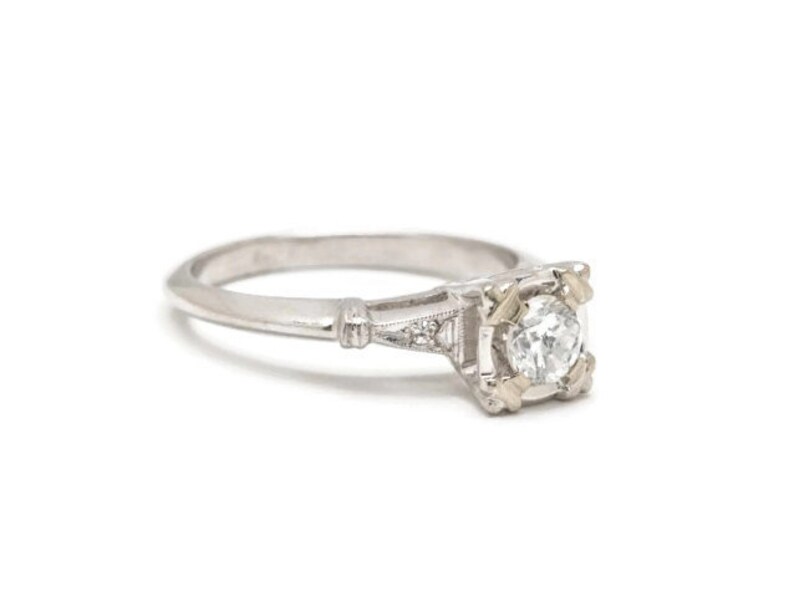 May include: A silver engagement ring with a single round diamond set in a square-shaped prong setting. The ring has a delicate, intricate design with small diamonds on the band.