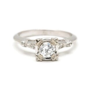 May include: A silver engagement ring with a round, clear diamond center stone. The diamond is set in a square frame with smaller diamonds on the band.