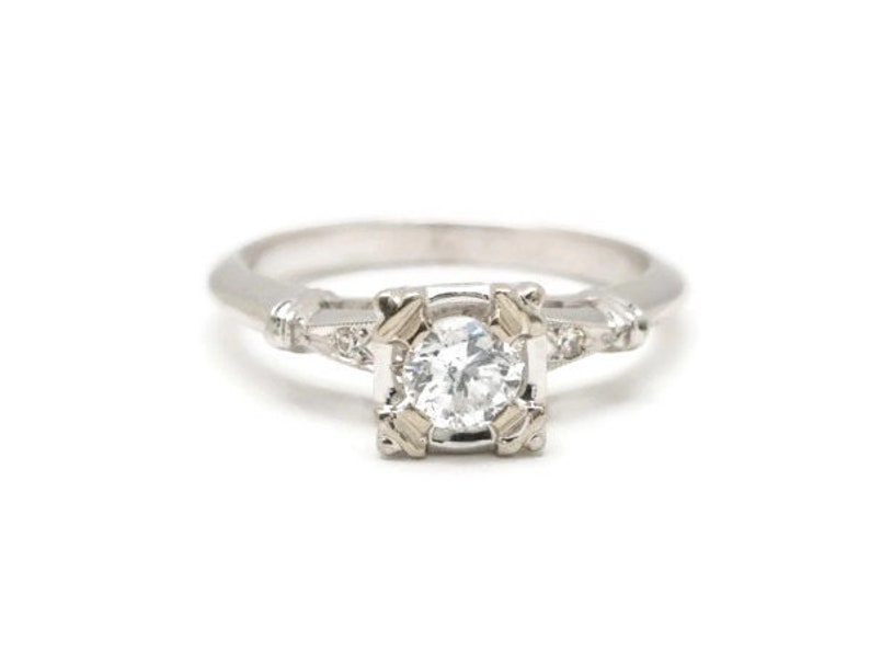 May include: A silver engagement ring with a round, clear diamond center stone. The diamond is set in a square frame with smaller diamonds on the band.