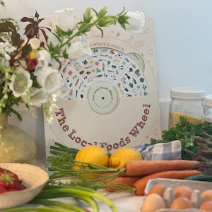May include: A white and red illustrated guide called "The Local Foods Wheel" with a spinning wheel showing different foods in season. The guide is surrounded by fresh produce, including lemons, carrots, asparagus, and strawberries.