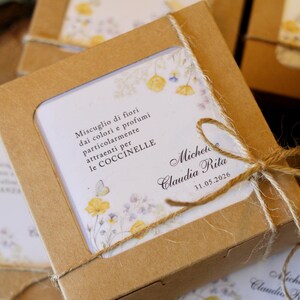 Può includere: Una scatola quadrata in cartone marrone con finestra trasparente, legata con spago naturale. All'interno, un cartoncino bianco presenta illustrazioni floreali gialle e viola, una piccola farfalla e il testo: Miscuglio di fiori dai colori e profumi particolarmente attraenti per le COCCINELLE, Michele, Claudia Rita, 31.05.2026.