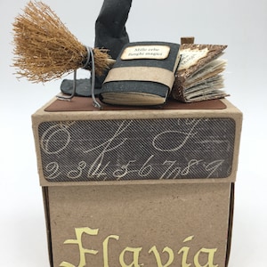 Può includere: Una scatola di cartone marrone con la parte superiore a motivi bianco e nero. La scatola ha un'iscrizione dorata che recita "Flavia". Sopra la scatola c'è un cappello da strega nero, una scopa marrone e una pila di libri con il testo "Mille erbe funghi magici".