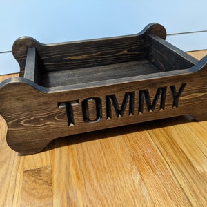 May include: A dark brown wooden dog bed shaped like a bone with the name "TOMMY" carved into the front.