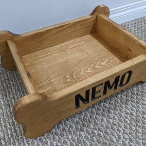May include: A wooden dog bed shaped like a bone with the name "NEMO" engraved on the side. The bed is made of natural wood and has a light brown stain.