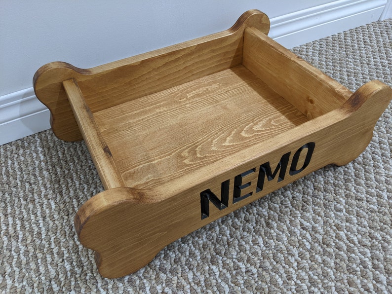 May include: A wooden dog bed shaped like a bone with the name "NEMO" engraved on the side. The bed is made of natural wood and has a light brown stain.