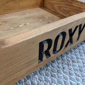 May include: A wooden dog bed with the name "ROXY" carved into the side. The bed is made of natural wood and has a light brown finish.