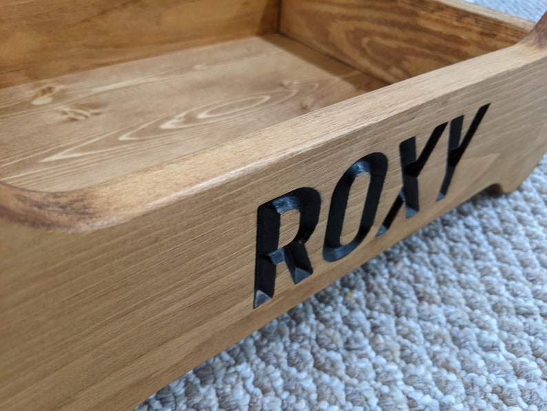 May include: A wooden dog bed with the name "ROXY" carved into the side. The bed is made of natural wood and has a light brown finish.