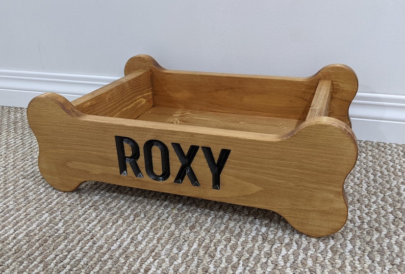 May include: A wooden dog bed shaped like a bone with the name "ROXY" engraved on the front. The bed is stained a light brown color.