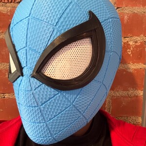 May include: A light blue Spider-Man mask with a textured surface and black eye frames. The mask has white mesh eye coverings. The mask is worn over a black shirt and a red jacket.