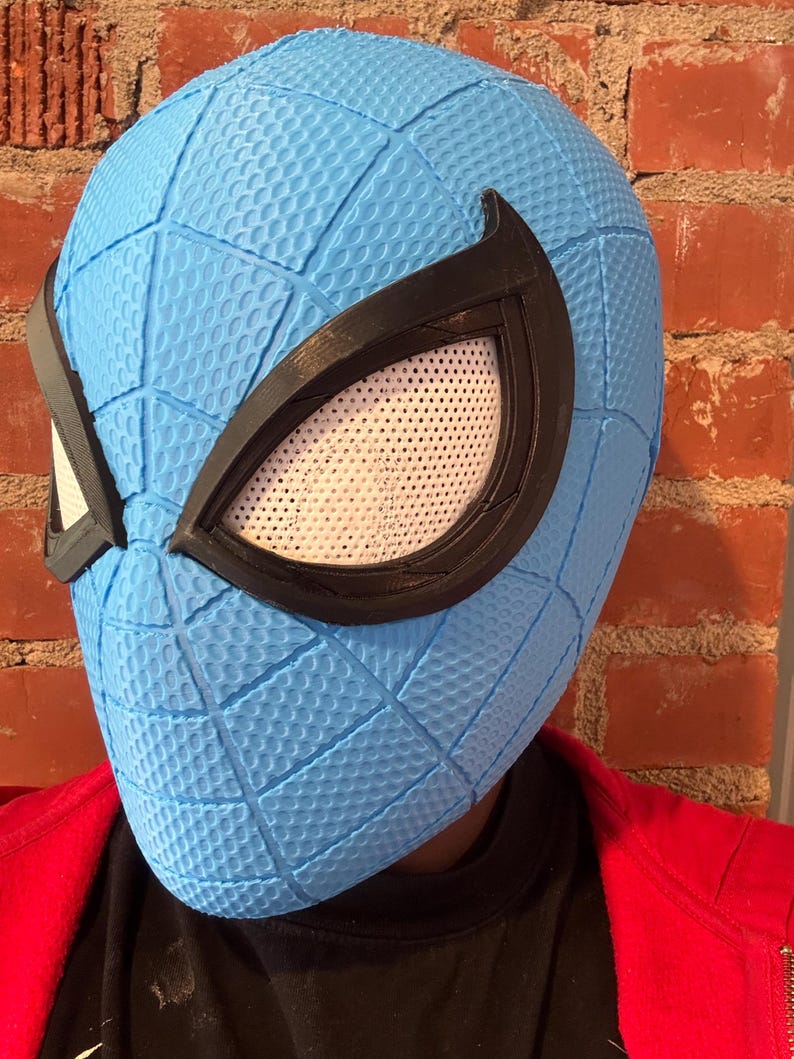 May include: A light blue Spider-Man mask with a textured surface and black eye frames. The mask has white mesh eye coverings. The mask is worn over a black shirt and a red jacket.