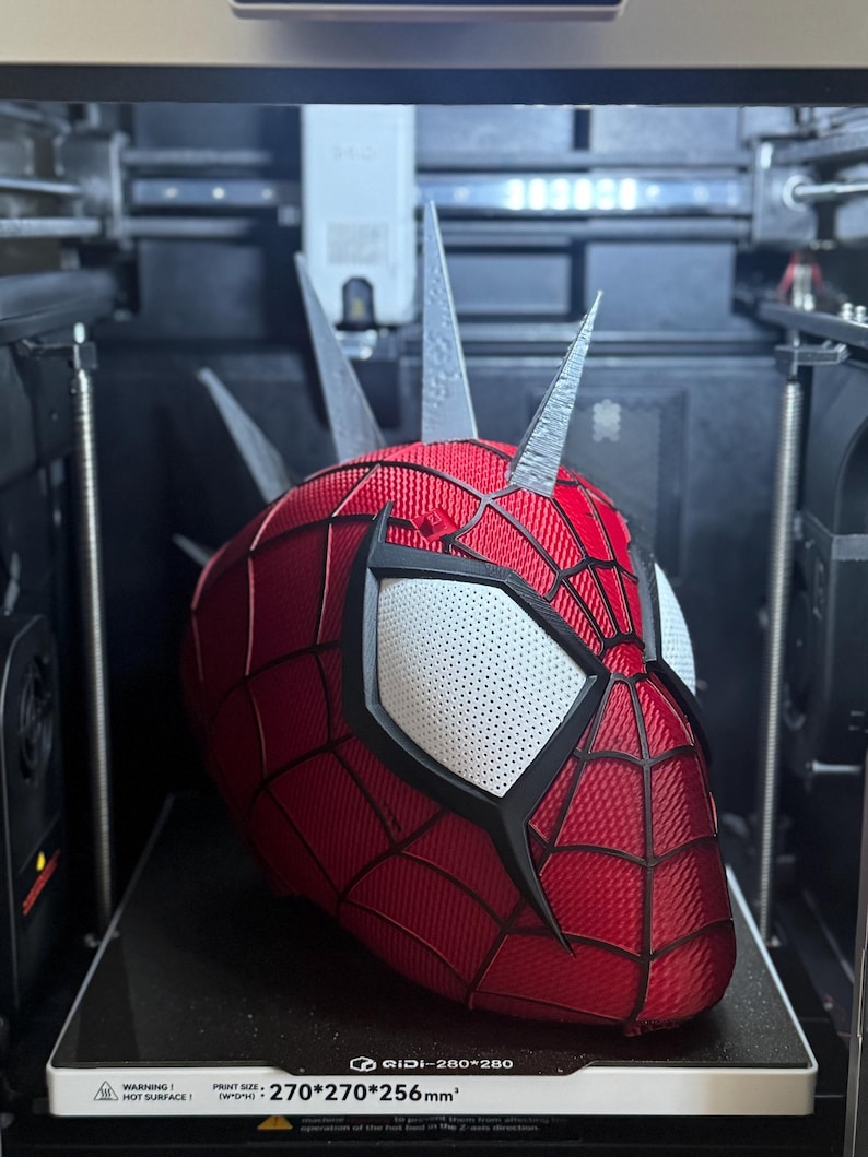 May include: A red and black Spider-Man mask with white eye lenses is being 3D printed. The mask has a webbed pattern and is in the process of being created inside a 3D printer. The printer bed has the text "GIDI-280*280" and "270*270*256mm&sup2;".