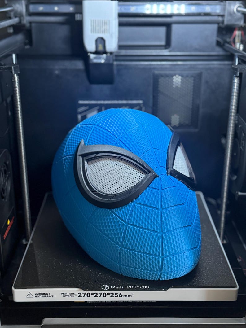May include: A blue Spider-Man helmet with black eye frames and a textured surface, 3D printed on a black platform. The print bed size is 270x270x256mm. The helmet is in a 3D printer.