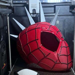 May include: A red and black 3D-printed Spider-Man helmet with silver spikes, featuring a web pattern design. The helmet is on a 3D printer bed, with a black and white accessory in the foreground. The printer has a warning about a hot surface.