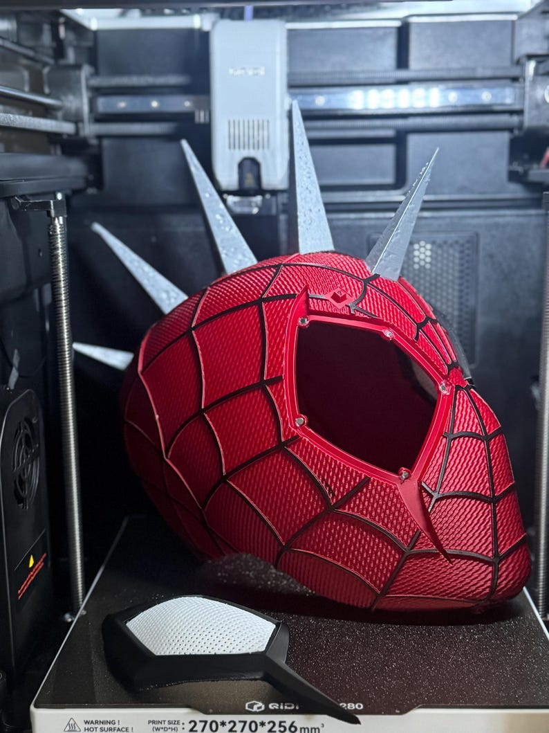 May include: A red and black 3D-printed Spider-Man helmet with silver spikes, featuring a web pattern design. The helmet is on a 3D printer bed, with a black and white accessory in the foreground. The printer has a warning about a hot surface.