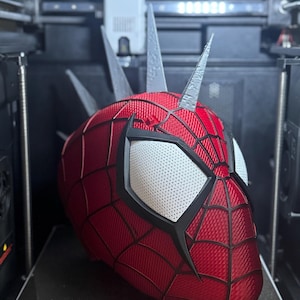 May include: A red and black Spider-Man mask with white eye lenses and silver spikes, 3D printed inside a 3D printer. The mask has a web pattern and is labeled with "QIDI-280*280" and "270*270*256mm&sup2;".