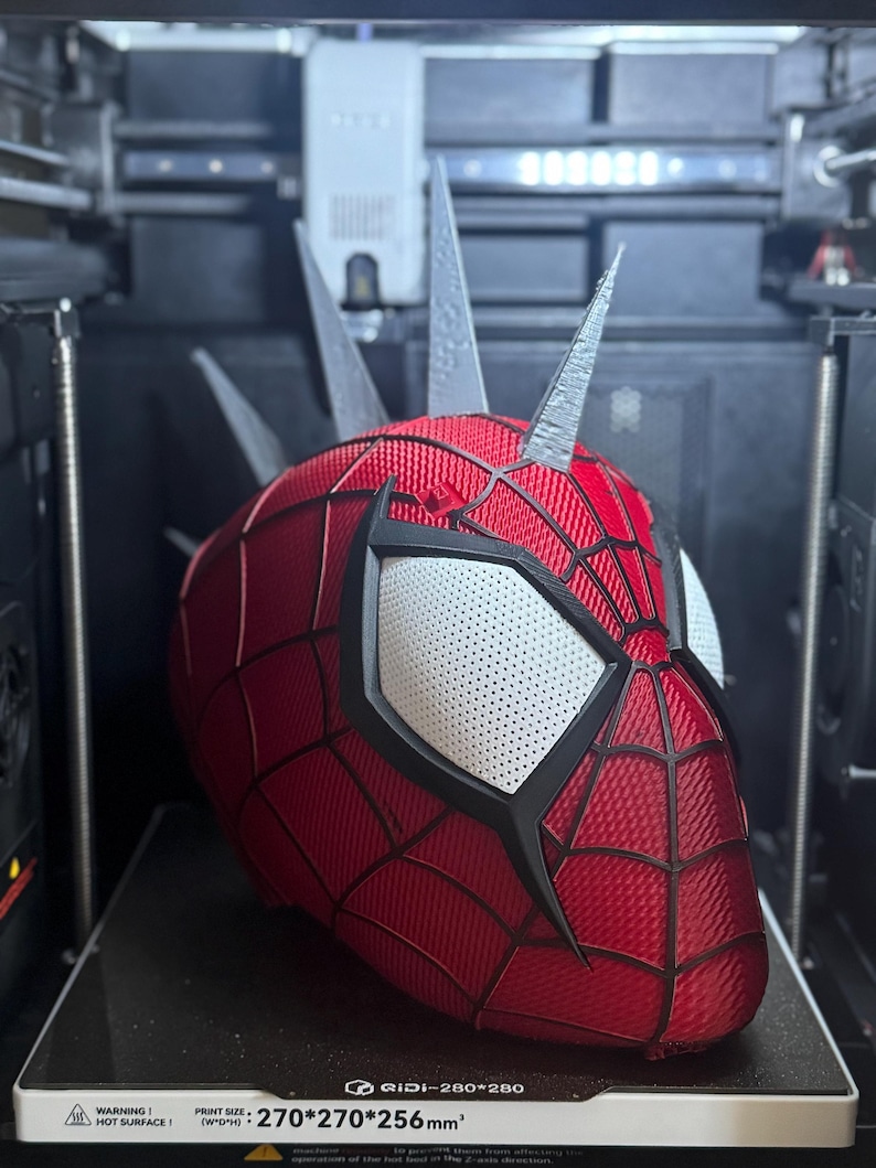 May include: A red and black Spider-Man mask with white eye lenses and silver spikes, 3D printed inside a 3D printer. The mask has a web pattern and is labeled with "QIDI-280*280" and "270*270*256mm&sup2;".