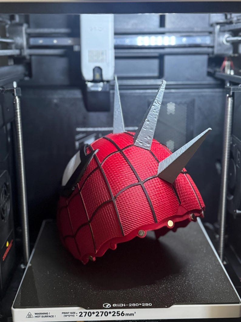 May include: A red and black Spider-Man mask with silver spikes is being 3D printed. The mask has a textured pattern and white eye details. The printer bed has the text "GIDI-280*280" and "270*270*256mm&sup3;".