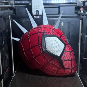 May include: A red and black Spider-Man mask with silver spikes is being 3D printed. The mask has white eye lenses and a textured surface. The printer bed has a warning about a hot surface and the print size is 270x270x256mm.