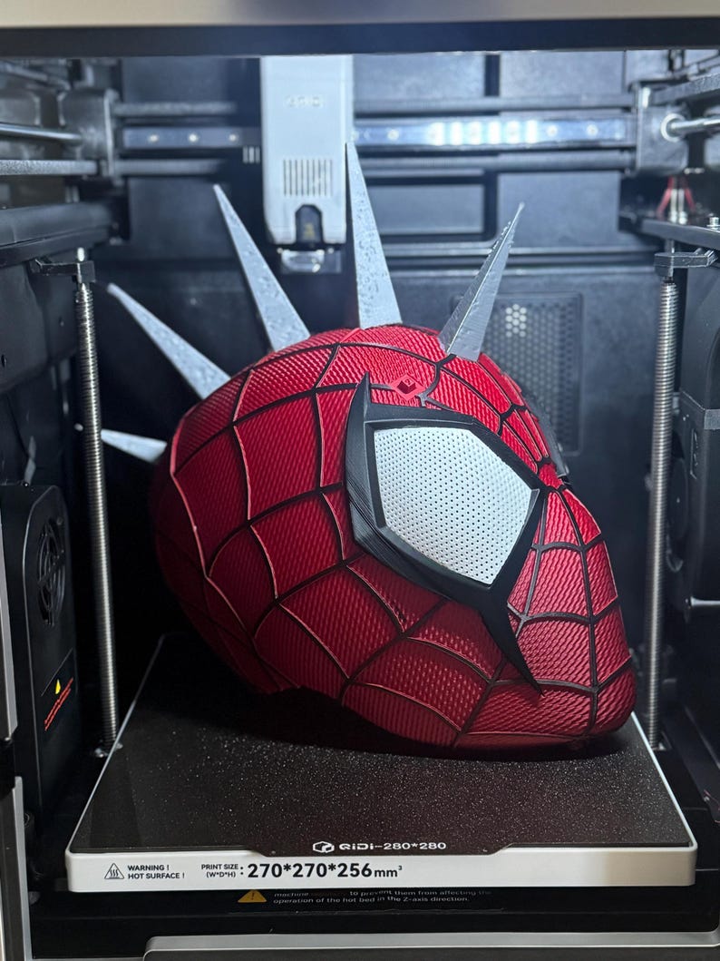 May include: A red and black Spider-Man mask with silver spikes is being 3D printed. The mask has white eye lenses and a textured surface. The printer bed has a warning about a hot surface and the print size is 270x270x256mm.
