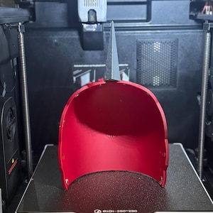 May include: A bright red, 3D-printed helmet with a silver, pointed top, resting on a 3D printer bed. The printer bed dimensions are 270mm x 270mm x 256mm. The helmet's interior is visible, and the printer's components are in the background.