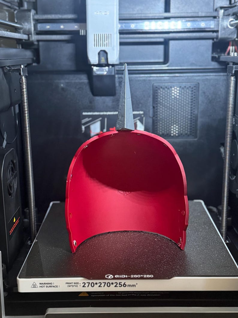 May include: A bright red, 3D-printed helmet with a silver, pointed top, resting on a 3D printer bed. The printer bed dimensions are 270mm x 270mm x 256mm. The helmet's interior is visible, and the printer's components are in the background.