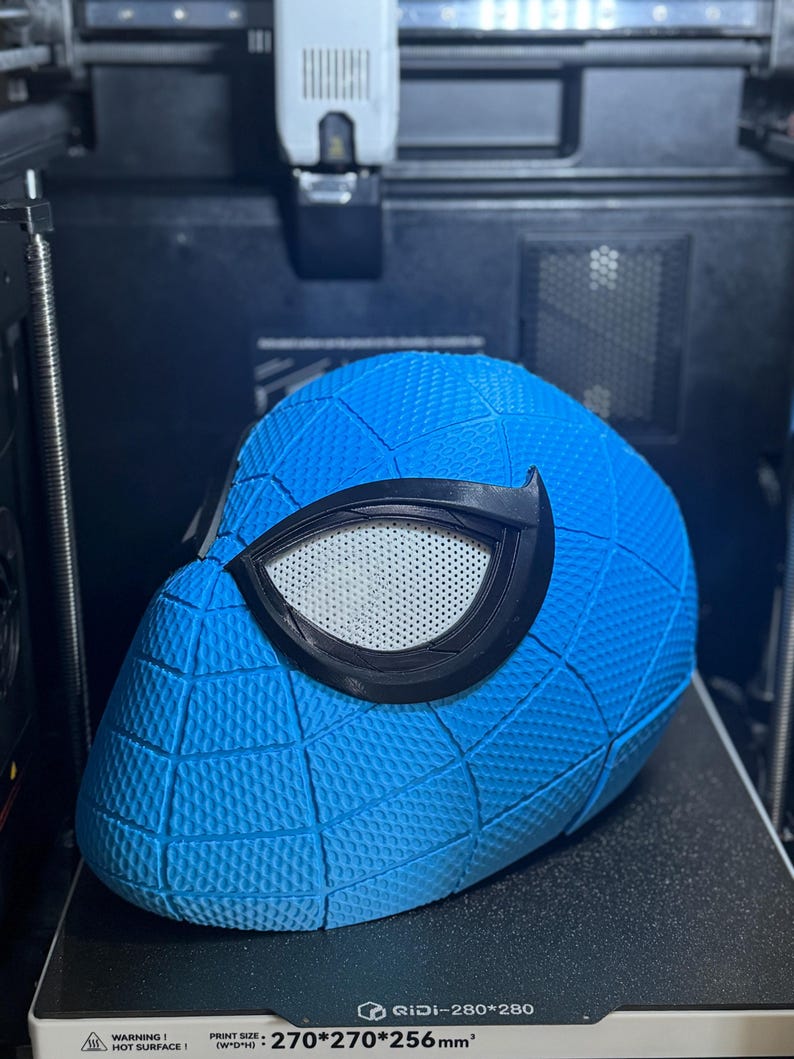 May include: A blue Spider-Man mask with a textured surface and black eye frames. The mask is on a 3D printer. The print size is 270x270x256mm.