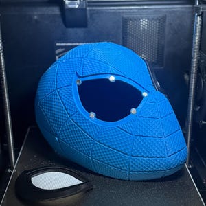 May include: A blue 3D-printed helmet with a textured surface and large eye openings. The helmet is on a 3D printer bed, with a separate eye lens piece. The print size is 270*270*256mm&sup3;.