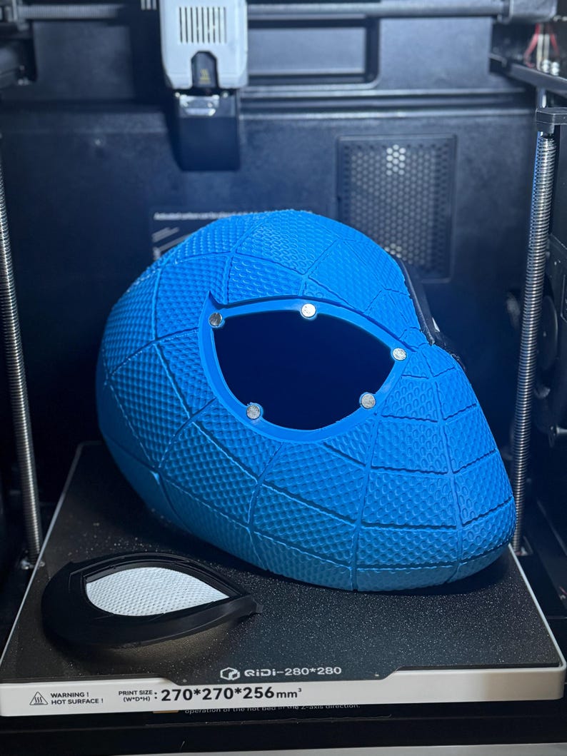 May include: A blue 3D-printed helmet with a textured surface and large eye openings. The helmet is on a 3D printer bed, with a separate eye lens piece. The print size is 270*270*256mm&sup3;.