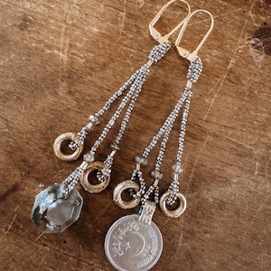 Puede incluir: Un par de pendientes de plata y oro con un dije de cristal y un dije de moneda de plata. La moneda tiene un diseño de media luna y estrella.