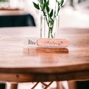 May include: A wooden and metal name card holder with the name "Marilyn" engraved on it. The holder is on a wooden table with a vase of green plants in the background.