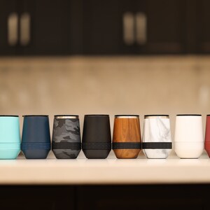 Puede incluir: Una fila de diez vasos aislados de acero inoxidable en varios colores y patrones, incluyendo rosa, azul, azul marino, camuflaje, negro, veta de madera, m&aacute;rmol, blanco y rojo.