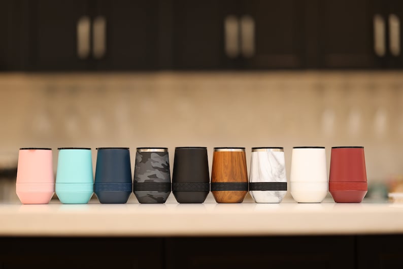 Puede incluir: Una fila de diez vasos aislados de acero inoxidable en varios colores y patrones, incluyendo rosa, azul, azul marino, camuflaje, negro, veta de madera, m&aacute;rmol, blanco y rojo.