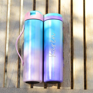 May include: Two stainless steel water bottles with a gradient design. The bottle on the left is blue and pink, while the bottle on the right is purple and pink. The bottle on the right has the name "Karen" engraved on it.