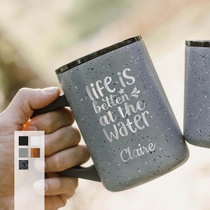 May include: A gray speckled travel mug with a black lid and a handle. The mug has the text "Life is better at the water" and the name "Claire" printed on it.