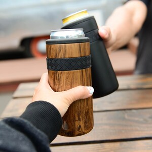 May include: Two people clinking insulated beverage holders. One holder is black and the other is wood-grain with a black band.