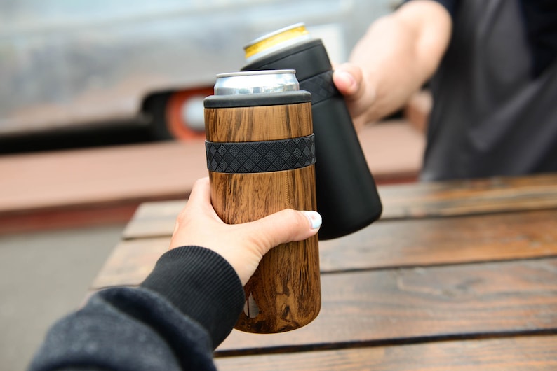 May include: Two people clinking insulated beverage holders. One holder is black and the other is wood-grain with a black band.