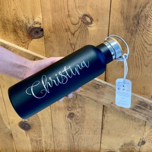 May include: Black stainless steel water bottle with a wooden lid and a carrying loop. The bottle is engraved with the name "Christina".