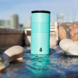 May include: A turquoise stainless steel water bottle with a black lid and a black logo. The bottle is sitting in a stream of water.