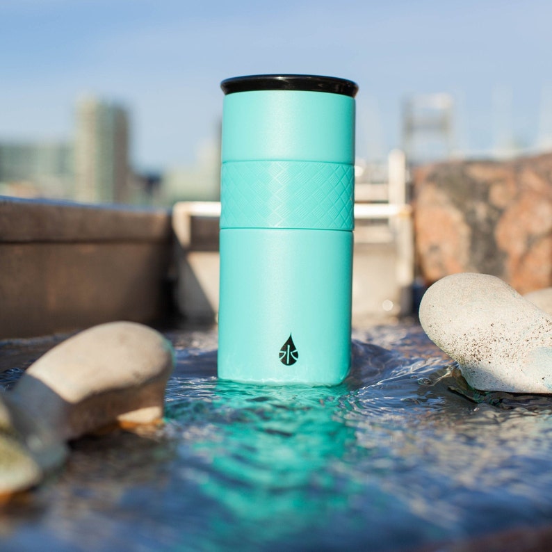May include: A turquoise stainless steel water bottle with a black lid and a black logo. The bottle is sitting in a stream of water.