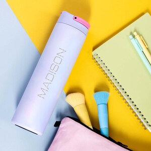 May include: A light purple stainless steel water bottle with a pink lid and the word "MADISON" engraved on the side. The bottle is on a blue and yellow surface with a notebook, makeup brushes, and a pink pouch.