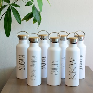 May include: Seven white water bottles with wooden lids and silver caps. Each bottle has a name printed on it: Susan, Chloe Happy, Nick, Ashley, Natalie, KK W, and Tracey.