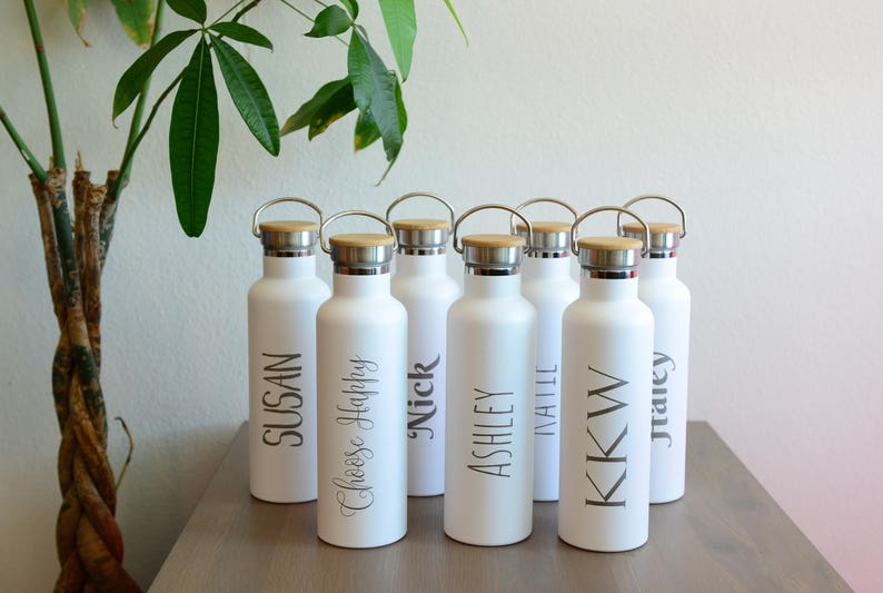 May include: Seven white water bottles with wooden lids and silver caps. Each bottle has a name printed on it: Susan, Chloe Happy, Nick, Ashley, Natalie, KK W, and Tracey.