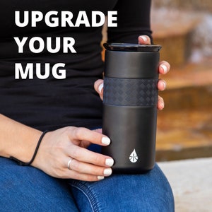 May include: A black travel mug with a textured black band around the middle. The mug has a white logo of a water drop on the bottom. The text "Upgrade Your Mug" is in white letters on a black background.
