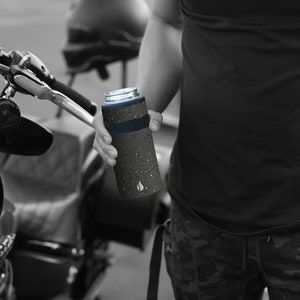 May include: A black and gray insulated can cooler with a speckled pattern and a blue band around the top. The cooler is being held by a person's hand.