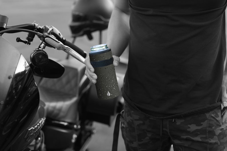 May include: A black and gray insulated can cooler with a speckled pattern and a blue band around the top. The cooler is being held by a person's hand.